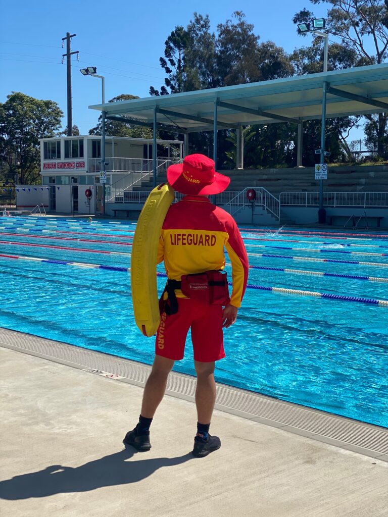 Photo of lifeguard standing looking out over an outdoor swimming pool.
